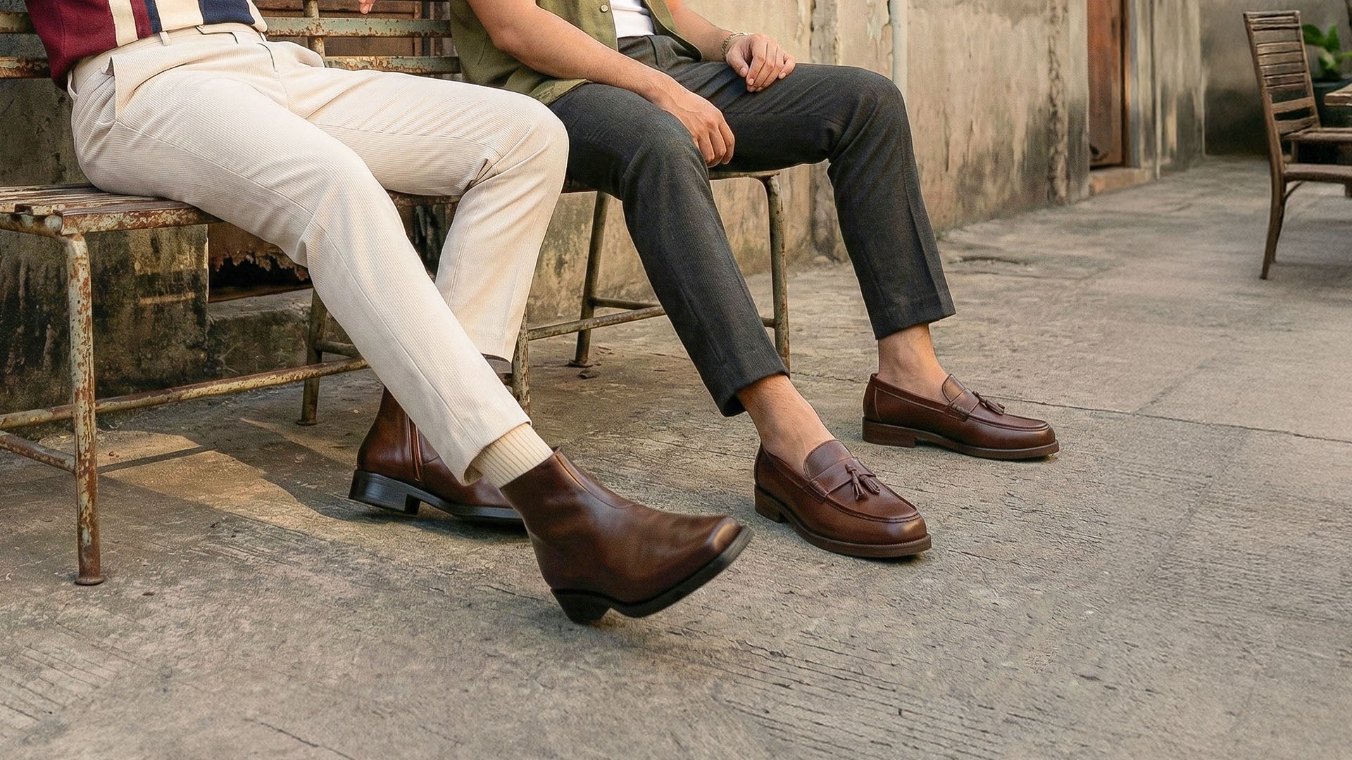 Two people sitting on a bench wearing brown leather chelsea and loafers.