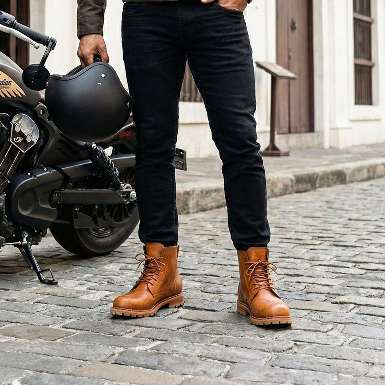 Person wearing brown leather boots standing next to a motorcycle on a cobblestone street.